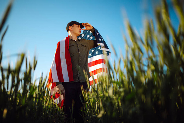 Ein junger Mann mit grünem Hemd und Mütze steht in die amerikanische Flagge gehüllt am grünen Weizenfeld. Patriotischer Junge feiert US-Unabhängigkeitstag am 4. Juli mit Nationalflagge in der Hand - Foto, Bild