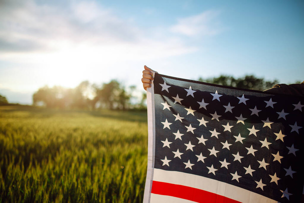 Young man wearing green shirt and cap lets the american flag fly on the wind at the green wheat field. Patriotic boy celebrates usa independence day on 4th of July with a national flag in his hands - Фото, изображение