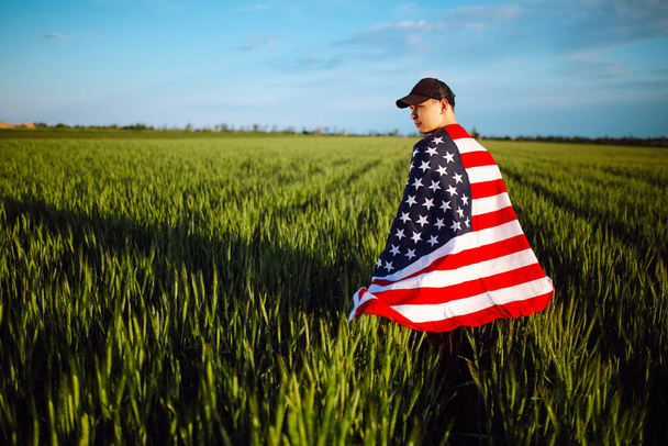 Ein junger Mann mit grünem Hemd und Mütze steht in die amerikanische Flagge gehüllt am grünen Weizenfeld. Patriotischer Junge feiert US-Unabhängigkeitstag am 4. Juli mit Nationalflagge in der Hand - Foto, Bild