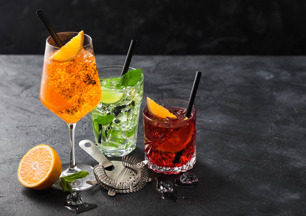 Glasses of spritz,mojito and negroni cocktails with ice cubes and lime and orange slices with mint leaf and black straw on dark background with strainer. Top view - Фото, изображение