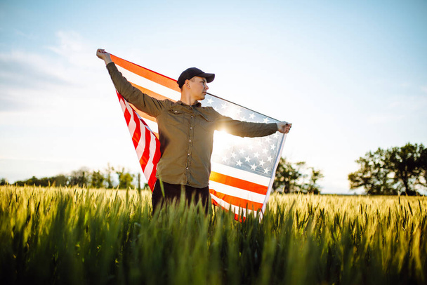 Young man wearing green shirt and cap lets the american flag fly on the wind at the green wheat field. Patriotic boy celebrates usa independence day on 4th of July with a national flag in his hands - Zdjęcie, obraz