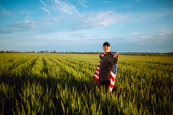 Young man wearing green shirt and cap stands wrapped in the american flag at the green wheat field. Patriotic boy celebrates usa independence day on the 4th of July with a national flag in his hands - Photo, Image