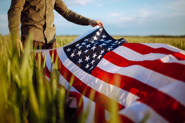 Young man wearing green shirt and cap lets the american flag fly on the wind at the green wheat field. Patriotic boy celebrates usa independence day on 4th of July with a national flag in his hands - Photo, Image