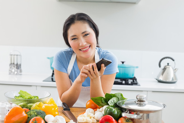 Smiling woman text messaging in front of vegetables in kitchen - Фото, изображение