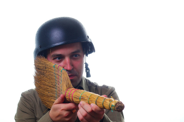 Muž v vojenské helmě s koštětem místo pušky na bílém pozadí. Vojenský humor. Koncept pohovka nebo lůžkové jednotky. - Fotografie, Obrázek