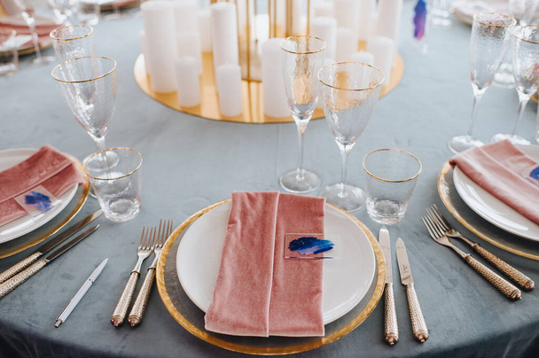 青いベルベットのテーブルクロスと宴会テーブル。プレートとカトラリー、バトラースメラルダグラス、ピンクベルベットのナプキン、キャンドル。バラの花、アジサイ、黄金の葉の構成で高いスタンド. - 写真・画像