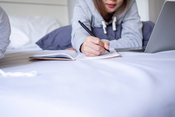 Liike-elämän naiset käyttävät rentoja vaatteita, jotka kirjoittavat kirjoja muistikirjaan. Työskennellä tai opiskella verkossa internet-yhteyden kautta tietokoneen sängyllä. Työn käsite kotoa tai opiskelusta kotona. - Valokuva, kuva