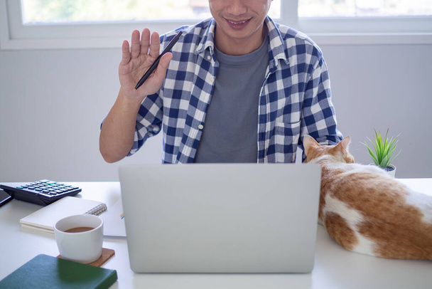 Азиатский бизнесмен машет рукой, чтобы поприветствовать своего партнера во время видеоконференции с рабочей группой дома
. - Фото, изображение