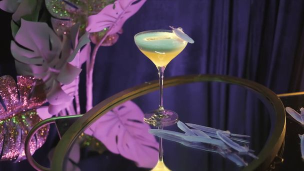 Um cocktail decorado com penas num restaurante. Imagens de stock. Close up de bebida alcoólica em vidro martini em pé sobre uma mesa em uma sala roxa com flores artificiais no fundo
. - Foto, Imagem
