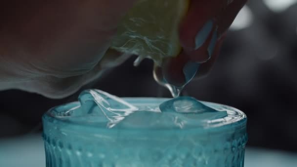 Close of female hand squeezing lemon juice into blue glass of beverage with ice cubes. Half of lemon fruit squeezing in slow motion macro view. - Materiał filmowy, wideo