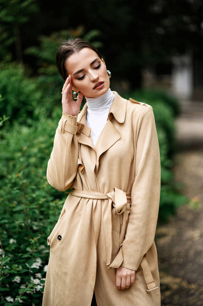 Beautiful brunette in a beige trench coat walking in the park - Foto, afbeelding