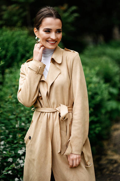 Beautiful brunette in a beige trench coat walking in the park - Foto, afbeelding