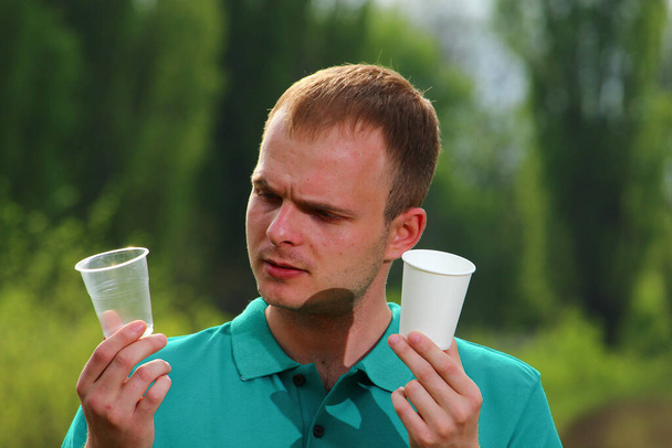 Marrs yeşil tişörtlü bir adam plastik bir tek kullanımlık bardak ve çevre dostu bir kağıt bardak gösteriyor.. - Fotoğraf, Görsel