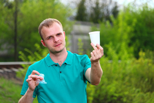 Ein junger Mann im grünen T-Shirt von Marrs hält einen zerknüllten Plastikbecher in der Hand und zeigt die richtige Alternative in Form eines Pappbechers.. - Foto, Bild