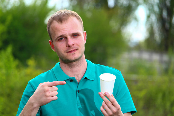 Ein Freiwilliger im grünen Marrs-T-Shirt zeigt seinen Finger auf einen Einwegbecher aus Papier. Das Konzept der richtigen ökologischen Wahl für Picknicks. - Foto, Bild