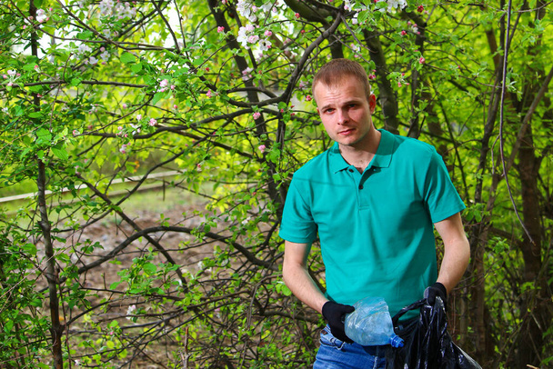 Volunteer young man in a Marrs green t-shirt with a garbage bag collects plastic non-decomposable garbage in nature. - Photo, image
