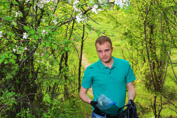 Молодой доброволец в зеленой футболке с мусорным мешком собирает пластиковый неразлагаемый мусор в природе
. - Фото, изображение