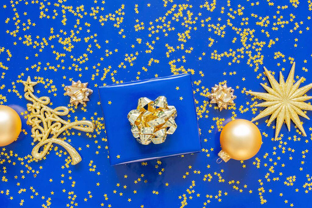 Κουτιά δώρων με ένα χρυσό τόξο και έλατο με μπάλες Χριστουγέννων σε μπλε φόντο, χρυσά λαμπερά αστέρια glitter σε μπλε φόντο, Χριστούγεννα έννοια, επίπεδη lay, top view - Φωτογραφία, εικόνα