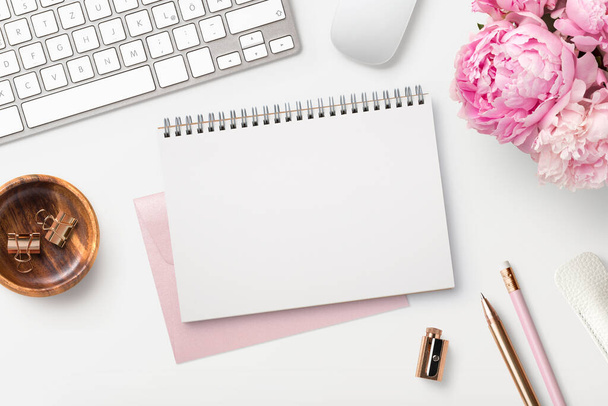 kobieca przestrzeń robocza / biurko z pustym otwartym notatnikiem, klawiaturą, stylowymi materiałami biurowymi / pisarskimi i różowymi piwoniami na białym tle, widok z góry - Zdjęcie, obraz