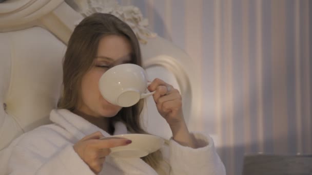 Girl Slavic appearance in white coat sitting on bed in hotel, drinking morning coffee, tea and showing emotions. Female smile. Close-up of young woman - Záběry, video