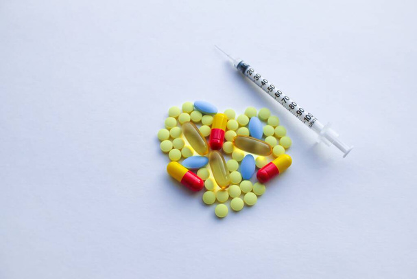 A lot of colorful tablets are laid out in the form of a heart, there is an insulin syringe on the background of a white textured rough sheet of paper with space for text - Foto, afbeelding