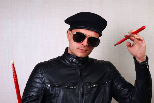 Real brutal rocker drummer with sticks dressed in black clothes: leather jacket, beret and sunglasses, lifestyle - Photo, Image