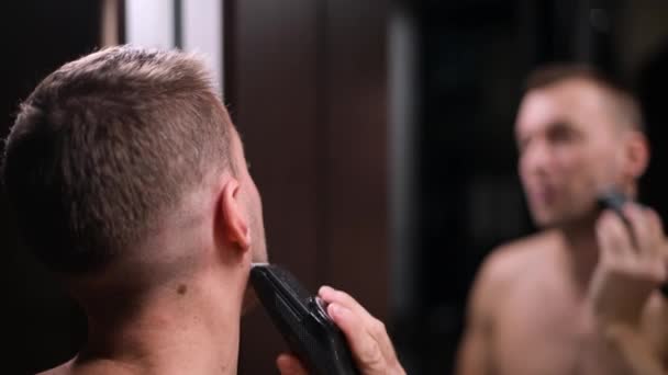 side view of a man cutting his beard with a trimmer in front of a mirror - Felvétel, videó