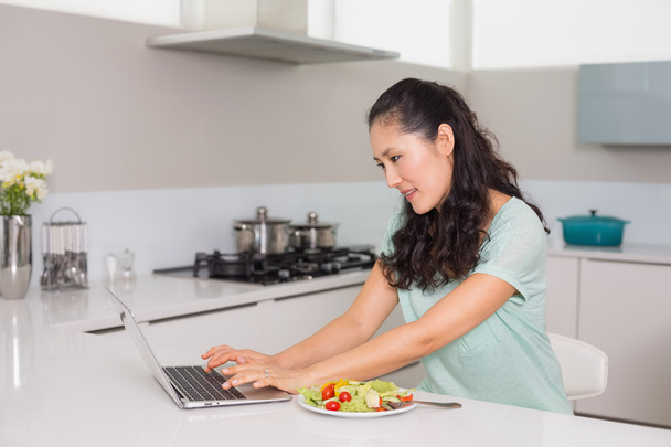 Young woman using laptop while having salad in kitchen - Valokuva, kuva