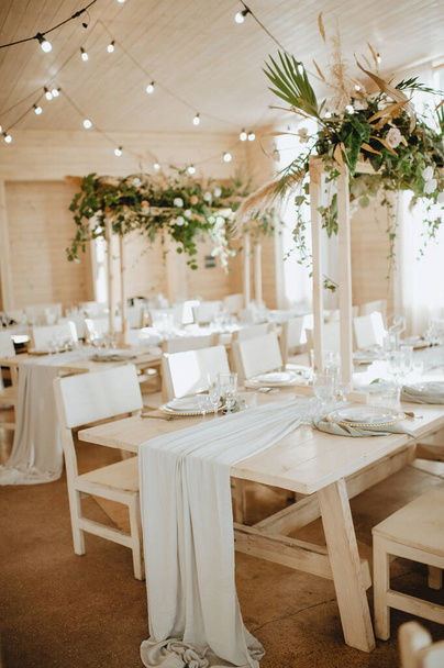 Restaurant, served table, rustic, huge compositions of flowers and tropical leaves on the table, plates, cutlery, crystal glasses, wooden chairs, garlands of light bulbs. - Fotó, kép