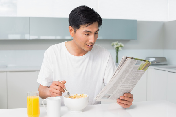 Young man having cereals while reading newspaper in kitchen - Photo, Image