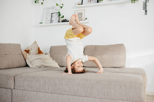 Beautiful little toddler standing on a head on sofa at home. Child Learn To Tumble. Tumbling Tutorial. Gymnastics at Home, Tricks - Fotó, kép