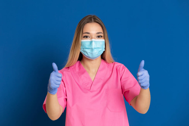 Young doctor wearing a mask face and a pink uniform on a blue background - Valokuva, kuva