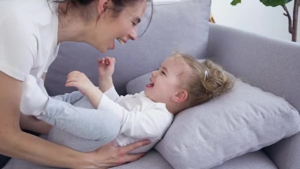 Retrato atraente de esplêndida jovem alegre e espirituosa que faz cócegas em sua linda filha de 1-2 anos que está deitada no sofá e rindo
 - Filmagem, Vídeo
