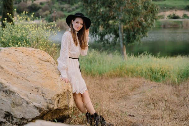 Young adorable girl in white dress and black hat, in spring, posing for camera. female portrait outdoor in mountain lake. Green fresh grass. Young unusual person enjoying nature - Photo, image