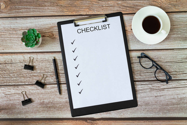 Checklist on blank page with pen, green plant, cup of coffee, eyeglasses and paper clips top view on wooden office or home table - Photo, Image
