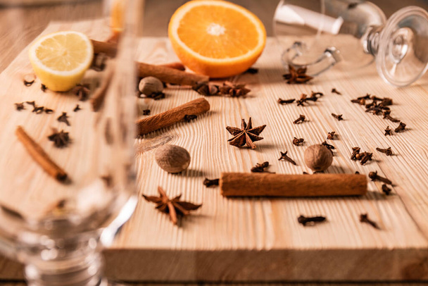Differential focus. Transparent empty glasses for mulled wine, fragrant cinnamon sticks,anise flowers stars, nutmegs, cloves, lemon and orange on structured natural wooden board, close up front view. - Fotoğraf, Görsel