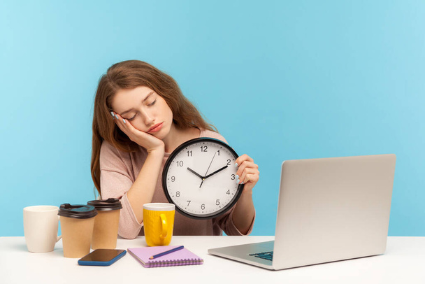Vyčerpaný kancelářský zaměstnanec pracuje dlouho do noci. Unavená žena obklopená šálky na kávu, spící a držící hodiny, pocit únavy, stres z práce přesčas. vnitřní studio záběr izolované na modrém pozadí - Fotografie, Obrázek
