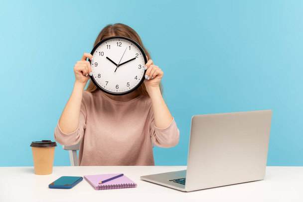 Time to work! Woman office employee sitting at workplace and covering face with big clock, concept of time management, schedule and business meeting appointment. indoor studio shot, blue background - Photo, image