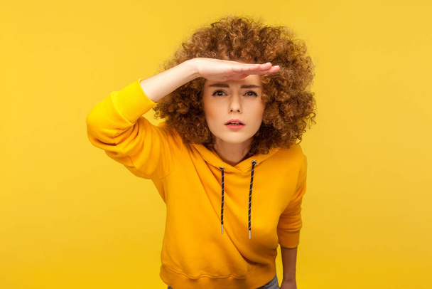 Portrait of curious curly-haired woman in urban style hoodie holding hand above eyes and peering into distance, looking far away, expecting and searching someone on horizon. studio shot isolated - Photo, image