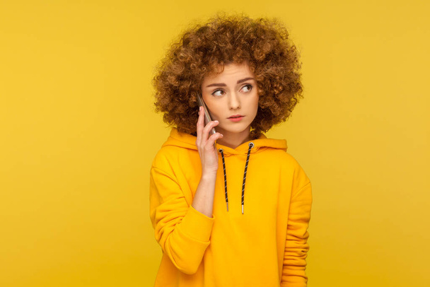 Portrait of upset curly-haired woman in urban style hoodie calling friend, talking on smartphone with serious unhappy expression, discussing bad news. indoor studio shot isolated on yellow background - 写真・画像