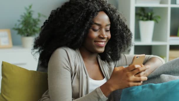 Beautiful curly African American young woman chatting and laughing on her smart phone. Living room. Portrait - Filmmaterial, Video