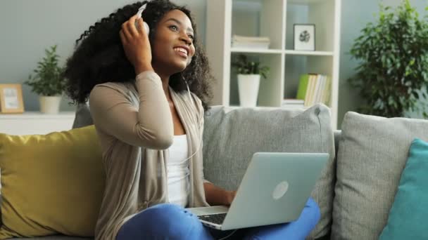 Pretty young curly African American woman in the white headphones listening to the music, dancing and working on her laptop while sitting on the sofa. Indoor. - Imágenes, Vídeo