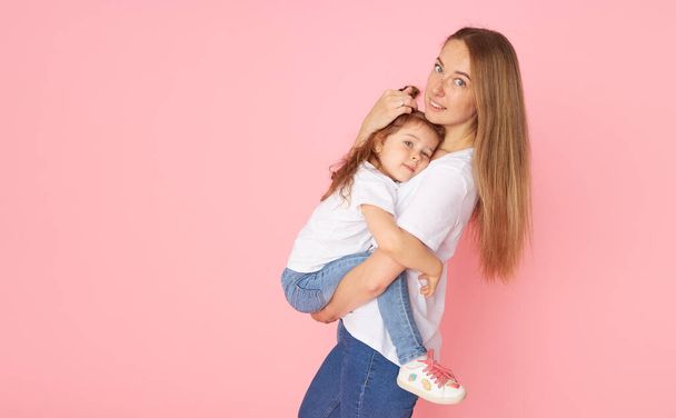 Mom and daughter in white t-shirts and jeans play and hug on a pink background. Caring for loved ones. Happy motherhood - Zdjęcie, obraz