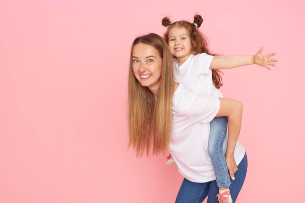 Mom and daughter in white t-shirts and jeans play and hug on a pink background. Caring for loved ones. Happy motherhood - Valokuva, kuva