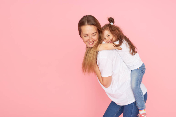 Mom and daughter in white t-shirts and jeans play and hug on a pink background. Caring for loved ones. Happy motherhood - Foto, Imagem