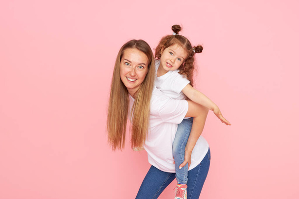 Mom and daughter in white t-shirts and jeans play and hug on a pink background. Caring for loved ones. Happy motherhood - Foto, Bild