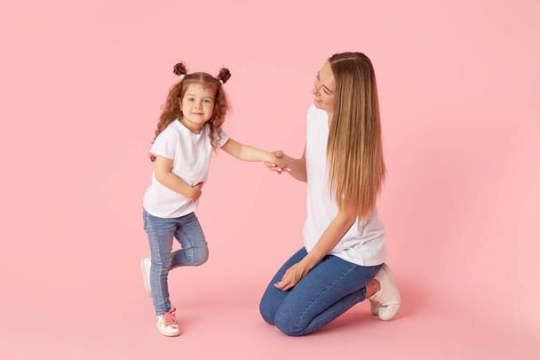 Mom and daughter in white t-shirts and jeans play and hug on a pink background. Caring for loved ones. Happy motherhood - Foto, immagini