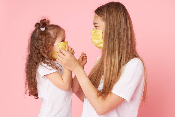Caring for loved ones. Young mother hugs little daughter in protective medical masks during Covid-19 pandemics. Studio pink background - Фото, изображение