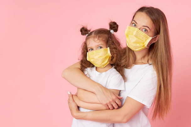 Caring for loved ones. Young mother hugs little daughter in protective medical masks during Covid-19 pandemics. Studio pink background - Zdjęcie, obraz