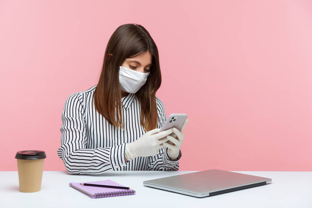 Online communication in self-isolation, coronavirus quarantine. Woman sitting safe healthy with hygienic face mask and protective gloves at workplace, typing message using mobile phone. studio shot - Фото, зображення